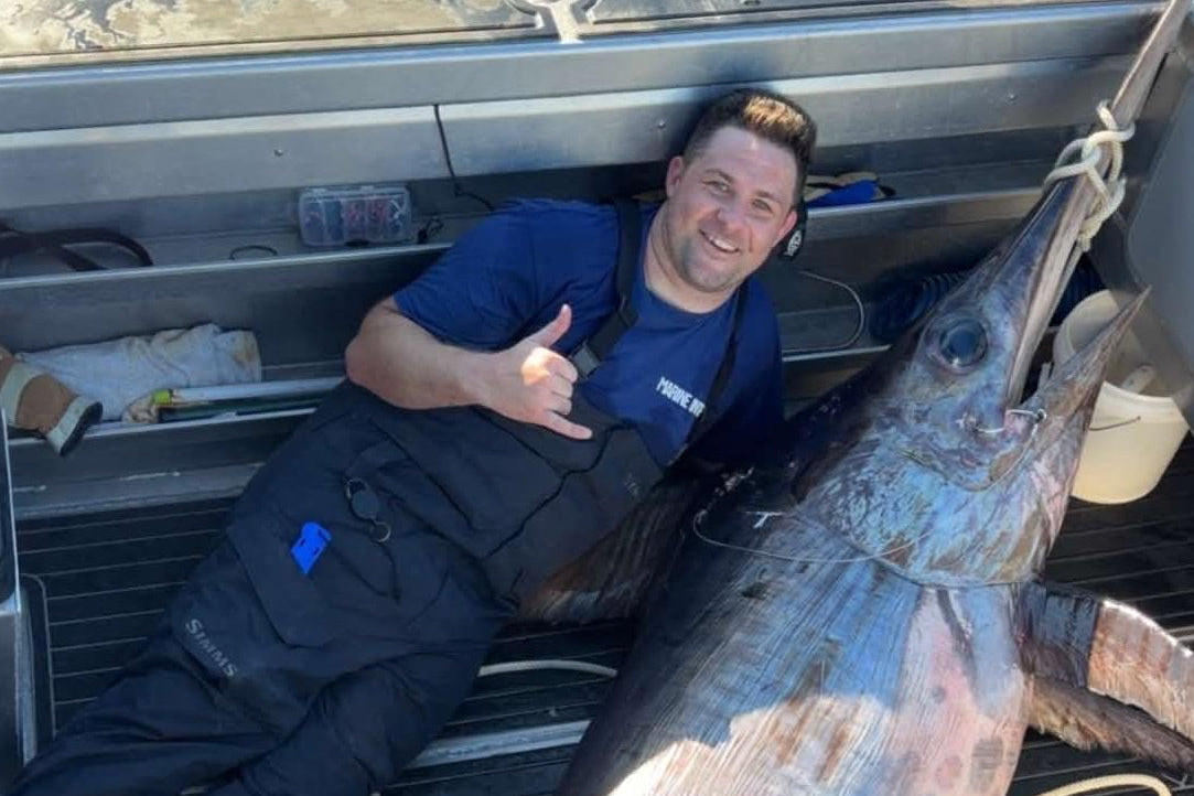 A man lying on the deck of a boat next to a very large sword fish, with water visible above and fishing gear around him.
